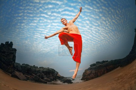 Un homme torse nu en pantalon rouge saute joyeusement contre un ciel dramatique, avec des falaises rocheuses et une plage de sable en contrebas, respirant un sentiment de liberté et d'aventure.