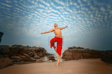 Un hombre sin camisa en pantalones rojos salta alegremente contra un cielo dramático, con acantilados rocosos y una playa de arena abajo, exudando una sensación de libertad y aventura.