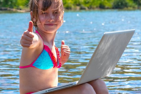 Jeune fille utilisant et apprenant avec ordinateur portable et montrant pouce vers le haut signe tout en voyageant montagnes et rivière. 