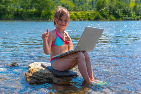 Young girl using and learning with laptop computer and showing thumbs up sign while traveling mountains and river. Horizontal image.