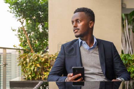 Téléchargez les photos : Portrait d'un bel homme d'affaires africain noir portant un costume tout en pensant et en utilisant le téléphone - en image libre de droit