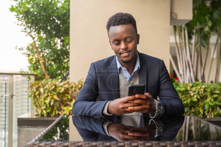 Téléchargez les photos : Portrait de beau noir homme d'affaires africain assis et utilisant le téléphone mobile à l'extérieur - en image libre de droit