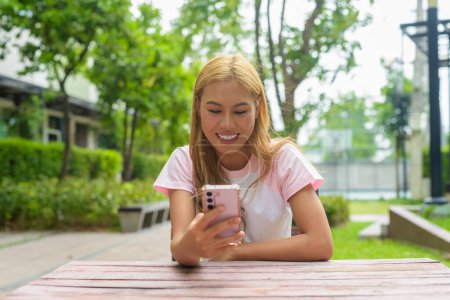 Photo for Portrait of beautiful happy Asian girl with blonde hair using phone outdoors while sitting - Royalty Free Image