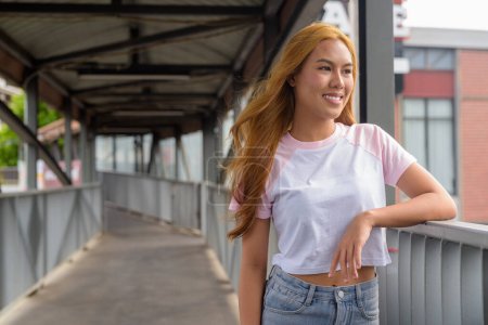 Photo for Portrait of beautiful happy Asian girl with blonde hair smiling and thinking outdoors - Royalty Free Image