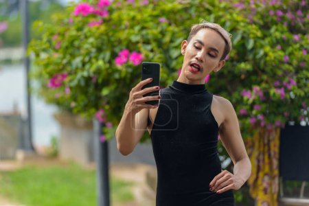 Beautiful Asian queer LGBT community supporter man with mustache wearing lipstick and jumpsuit using phone outdoors in park