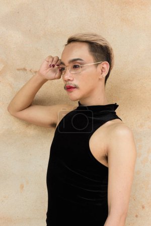 Retrato de hermoso asiático queer LGBT comunidad partidario hombre con bigote usando lápiz labial y mono contra fondo de pared beige