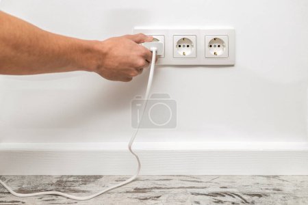 Photo for The human hand plugging a phone adapter in a triple white electrical outlet, situated on a white wall, front view. - Royalty Free Image