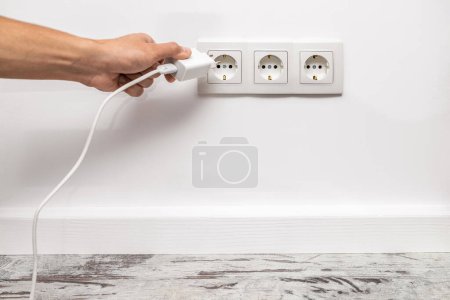 Photo for The human hand unplugging a phone adapter from a triple white electrical outlet, situated on a white wall. - Royalty Free Image