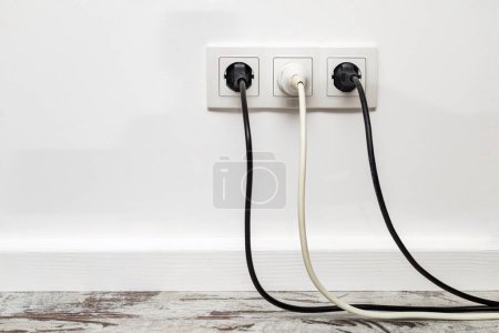 Photo for Triple white outlet installed on a white wall with two black plugs and a white plug. Front view - Royalty Free Image