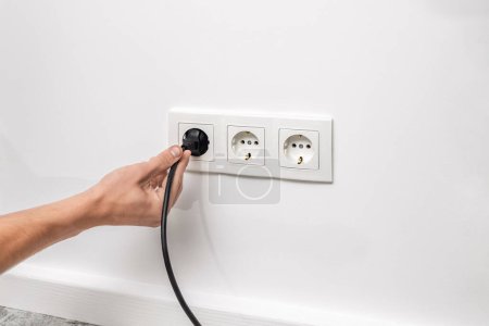 Photo for Human hand holding black cable plugged into triple white electrical outlet on the white wall - Royalty Free Image