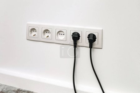 Photo for White five-outlet socket installed on white wall with two black plugs inserted, front view. - Royalty Free Image
