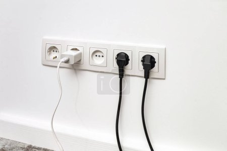 Photo for White five-way wall power socket installed on a white wall with two black plugs and a white phone adapter, front view. - Royalty Free Image