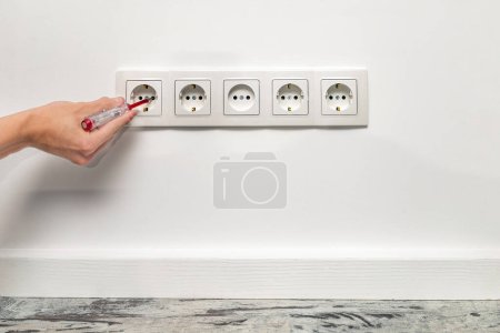 Photo for Human hand is holding a screwdriver plugged into an outlet. White five-way wall power socket installed on the white wall, front view. - Royalty Free Image