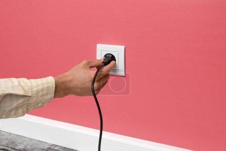 Photo for Human hand holding black cable plugged into white electrical outlet on the pink wall - Royalty Free Image