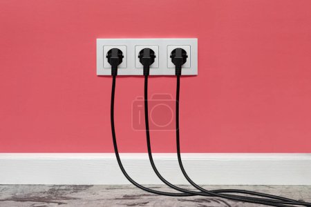 Photo for Triple white outlet installed on the pink wall with three black plugs inserted, front view. - Royalty Free Image