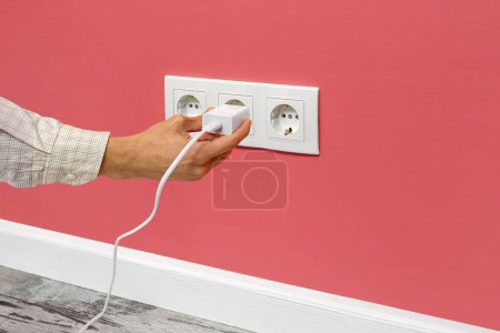 Photo for The human hand plugging a phone adapter in a triple white electrical outlet, situated on a pink wall, side view. - Royalty Free Image