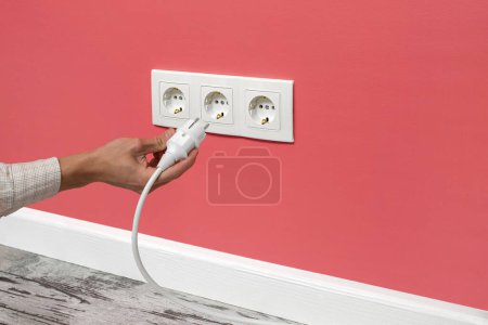 Photo for Human hand plugging white cable to triple white electrical outlet on the pink wall - Royalty Free Image