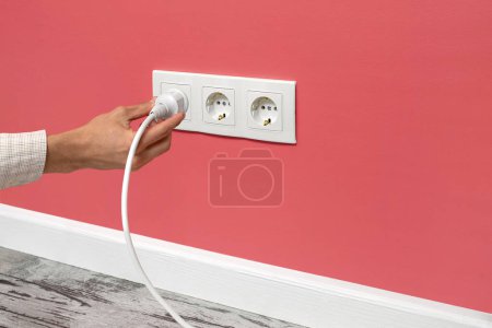 Photo for Human hand holding white cable plugged into triple white electrical outlet on the pink wall - Royalty Free Image