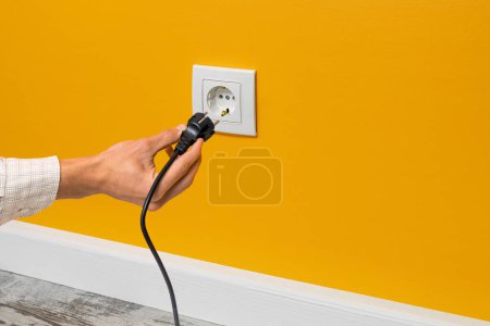 Photo for Human hand plugging black cable into white outlet on yellow wall - Royalty Free Image