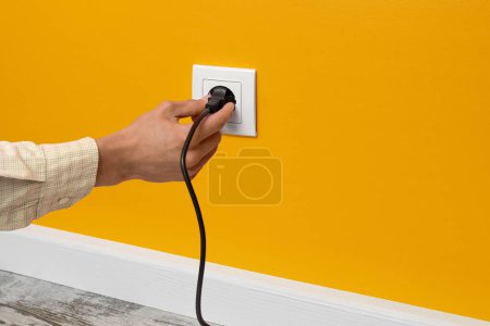 Photo for Human hand holding black cable plugged into white electrical outlet on the yellow wall - Royalty Free Image