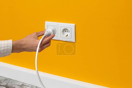 Photo for Human hand holding white cable plugged into double white electrical outlet on the yellow wall - Royalty Free Image