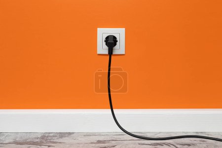 Photo for White outlet installed on orange wall with inserted black electrical plug, front view. - Royalty Free Image