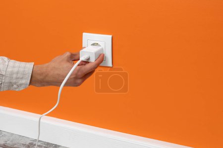 Photo for The human hand plugging a phone adapter in a white electrical outlet, situated on a orange wall, side view. - Royalty Free Image