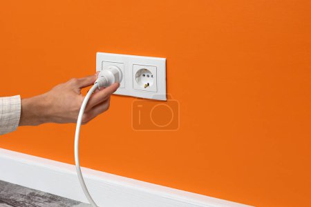 Photo for Human hand holding white cable plugged into double white electrical outlet on the orange wall - Royalty Free Image