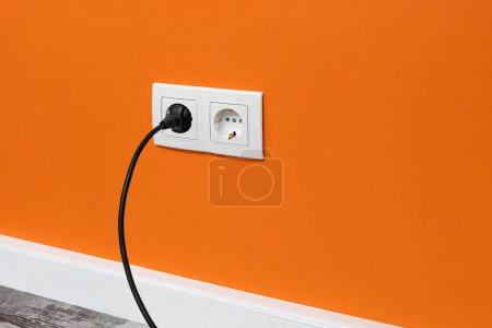 Photo for White double outlet installed on orange wall with inserted black electrical plug, side view. - Royalty Free Image
