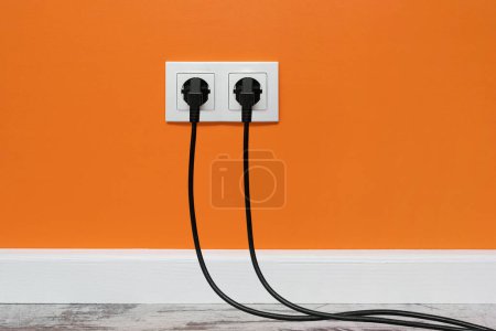 Photo for White double outlet installed on orange wall with two black plugs inserted, front view. - Royalty Free Image