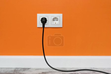 Photo for White double outlet installed on orange wall with inserted black electrical plug, front view. - Royalty Free Image