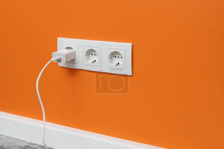 Photo for White triple outlet installed on the orange wall with inserted phone adapter, side view. - Royalty Free Image