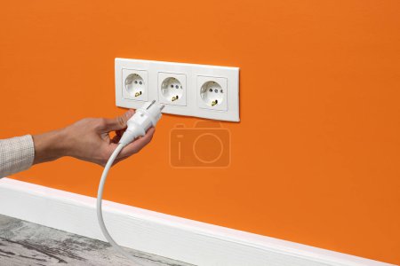 Photo for Human hand plugging white cable to triple white electrical outlet on the orange wall - Royalty Free Image