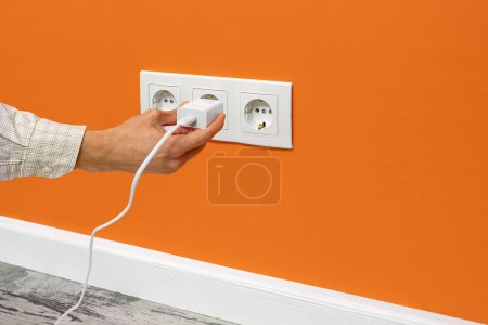 Photo for The human hand plugging a phone adapter in a triple white electrical outlet, situated on a orange wall, side view. - Royalty Free Image