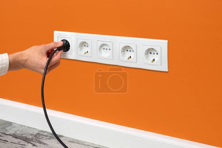 Photo for Human hand plugging black cable to a five-way wall power socket on the orange wall - Royalty Free Image