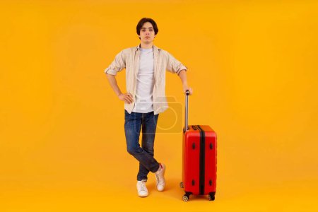Un viajero masculino con atuendo casual de pie con una maleta roja, absorto en su teléfono sobre un fondo amarillo.