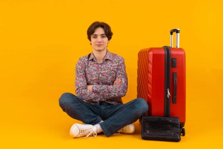 A male traveler in casual attire standing with a red rolling suitcase, engrossed in his phone on a yellow backdrop.