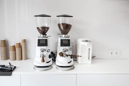 Téléchargez les photos : Deux moulins à café commerciaux blancs avec affichages numériques, prêts à l'emploi dans un cadre de café, flanqués de tasses en papier empilées et d'une bouilloire électrique blanche. - en image libre de droit