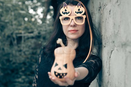 Téléchargez les photos : Gros plan portrait d'une jolie femme habillée en costume d'Halloween près d'un mur de pierre. sorcière aux cheveux orange vif, lunettes aux citrouilles, tient Jack-o-lanterne dans sa main. - en image libre de droit