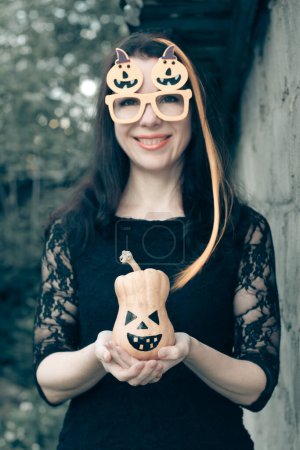 Téléchargez les photos : Gros plan portrait d'une jolie femme habillée en costume d'Halloween près d'un mur de pierre. sorcière aux cheveux orange vif, lunettes aux citrouilles, tient Jack-o-lanterne dans sa main. - en image libre de droit