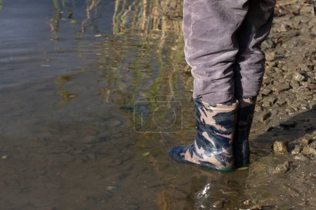 Kinderfüße in Gummistiefeln. Ein Kind in militärfarbenen Regenstiefeln steht am Ufer des Flusses im Wasser. Wasserdichte Kinderschuhe. Draußen