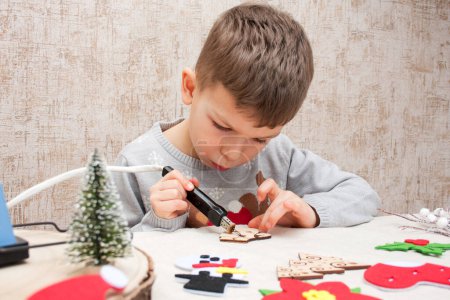 Photo for Preschooler boy burning an ornament on wooden blank with pyrograph, sitting at table at home. child is preparing for Christmas and New Year. Children creativity. - Royalty Free Image