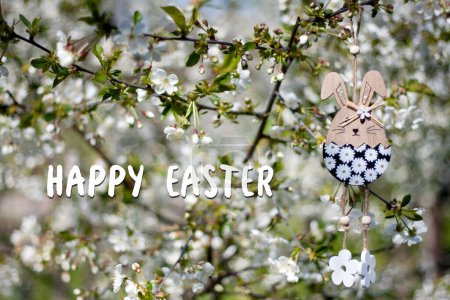 Tarjeta de felicitación con texto Feliz Pascua en inglés. Decoración de Pascua. Juguete decorativo de madera para conejito de Pascua en el fondo del floreciente jardín de primavera. Exterior