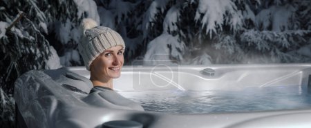 smiling woman with knitted hat relaxing in outdoor hot tub at snowy winter. banner with copy space