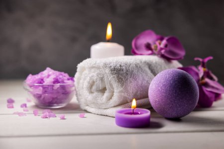 Foto de Bomba de baño púrpura, cristales de sal marina, toalla y velas perfumadas en la mesa de madera. cuidado de la piel corporal. spa de bienestar - Imagen libre de derechos