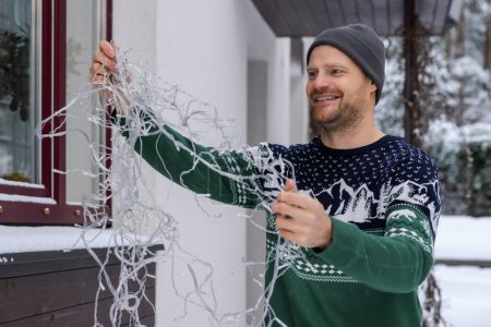 Foto de Hombre feliz al aire libre en el suéter de Navidad desenredar luces de cuerda para la decoración exterior de la casa - Imagen libre de derechos
