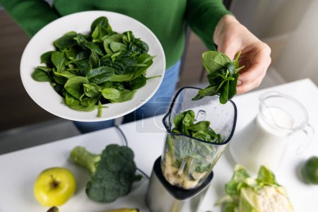 Foto de Mujer pone espinacas verdes frescas en la licuadora para hacer batido de desintoxicación saludable de verduras y frutas en la cocina casera - Imagen libre de derechos