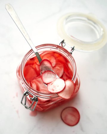 Jar of pickled thinly sliced pink radishes. A spoon is sticking out of the jar. White background. Korean dish. High quality photo