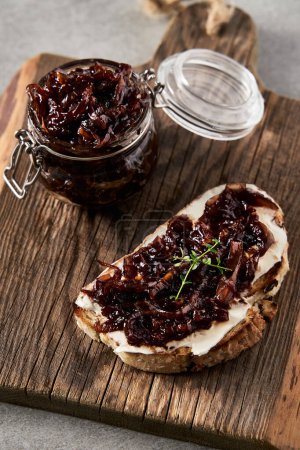 Delicious French Appetizer Freshly Cooked Onion Confit on toast with cream cheese and thyme. Wood board and jar with onion marmalade. Concrete loft background. . High quality photo