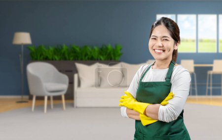 Téléchargez les photos : Asiatique femme de ménage porter des gants en caoutchouc jaune et tablier vert souriant dans le salon - en image libre de droit